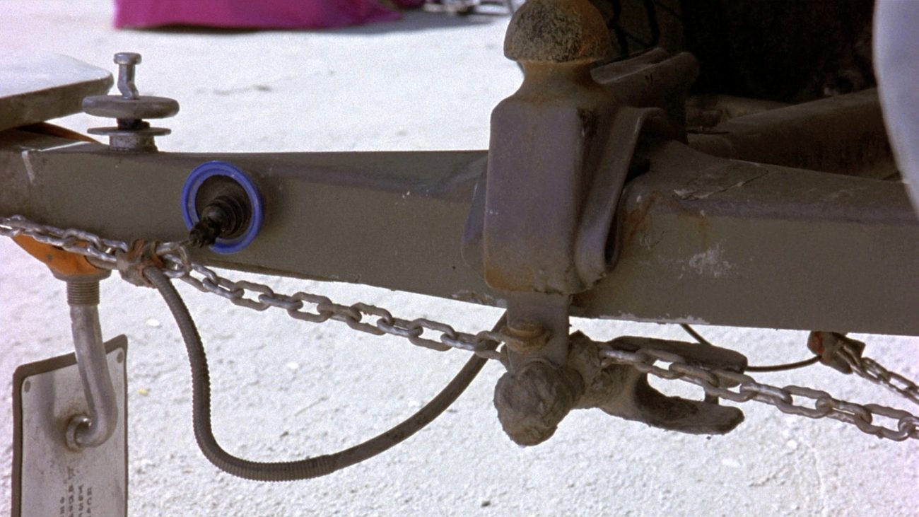 Close-up of a trailer tongue coupled to a truck: the coupler seated down over a greasy tow ball, safety chains crossed beneath it, a coiled electrical lead running back to the socket, the jack and a stamped rating plate at the left, all over pale gravel.