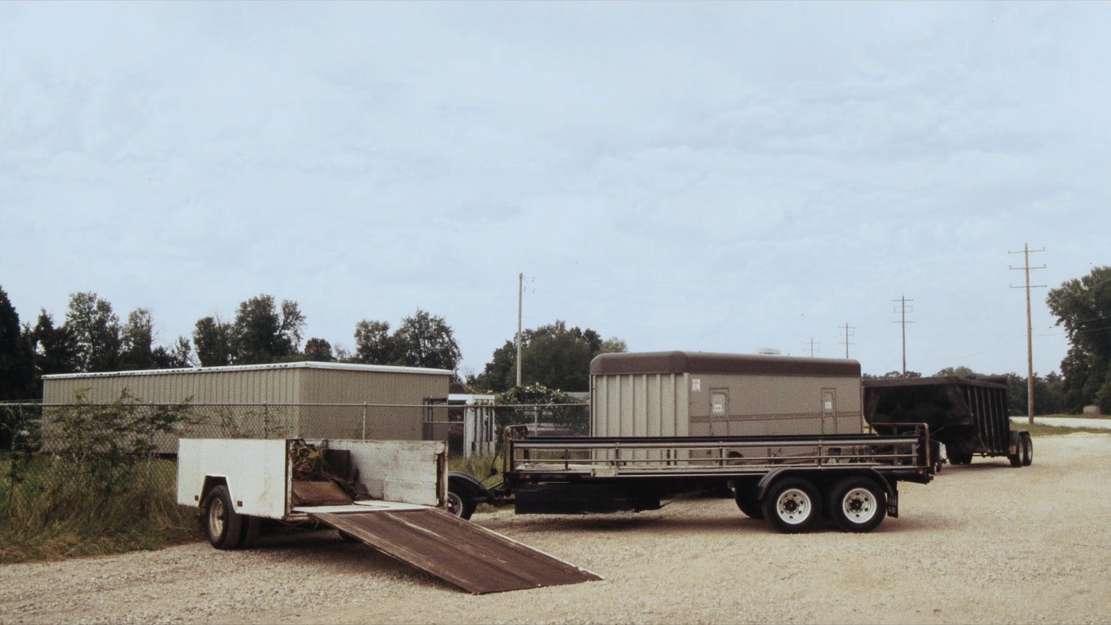 A gravel rental yard on an overcast day with a row of trailers parked in a line: a white single-axle utility trailer with its ramp down, a tandem-axle flatbed, and a dump trailer under a tarp, with a chain-link fence and a low metal shed behind.